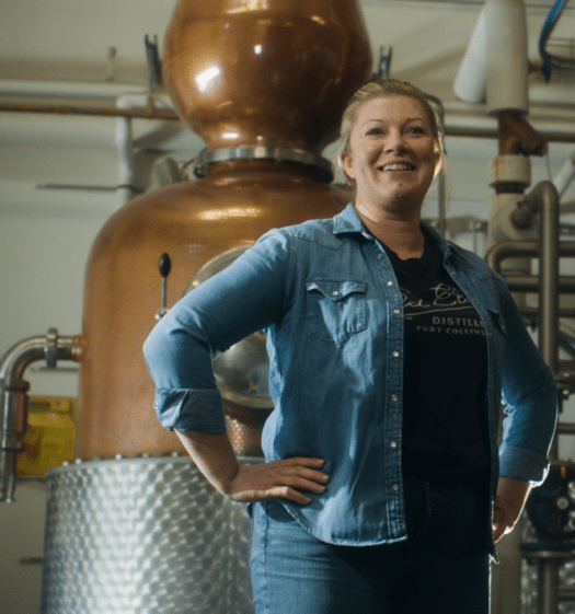 Melinda Maddox in a jean jacket standing in front of a whiskey still. 